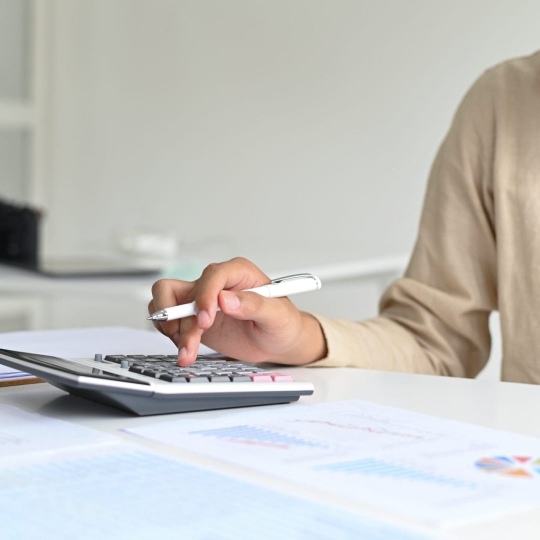 A young man with a pen in hand is operating a calculator, graphi A young man with a pen in hand is operating a calculator, graphing data and laptop on the desk.