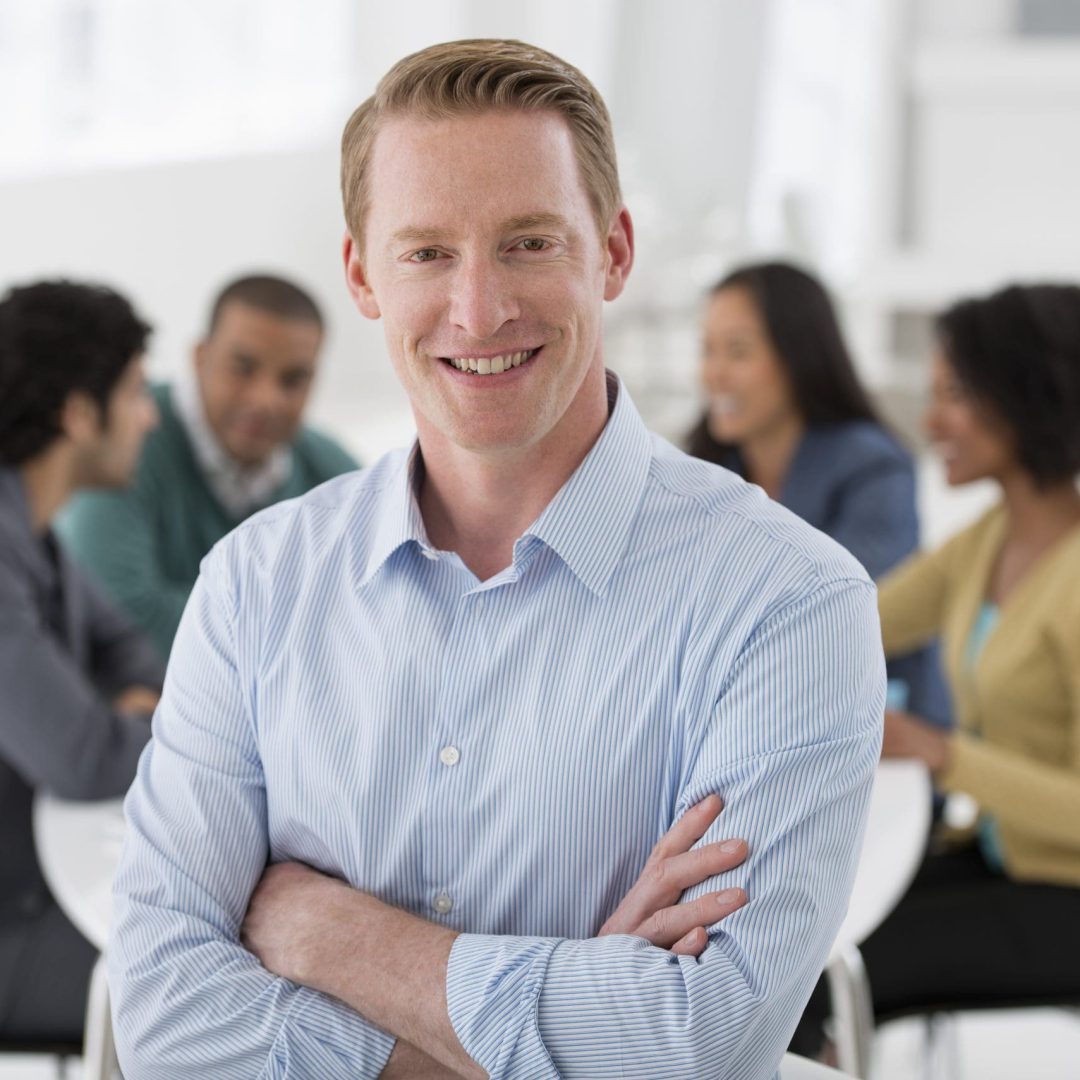 An office in the city. Business. Team meetings. A group sitting down around a table. One person in the foreground. A man smiling confidently.