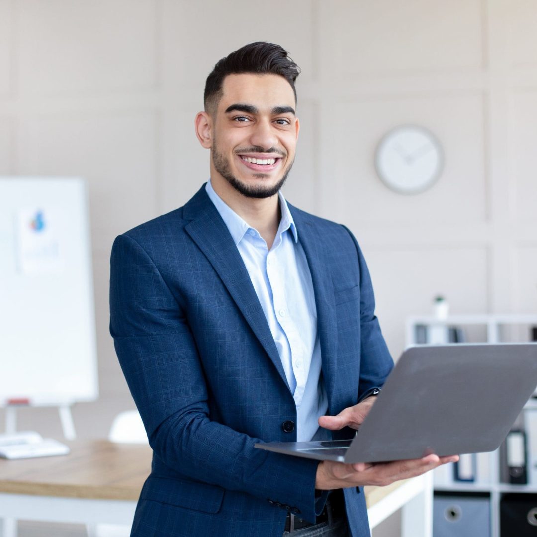 Cheerful Arab businessman in elegant suit holding laptop pc, smiling at camera in modern office. Young male employee conducting online company meeting or conference on computer