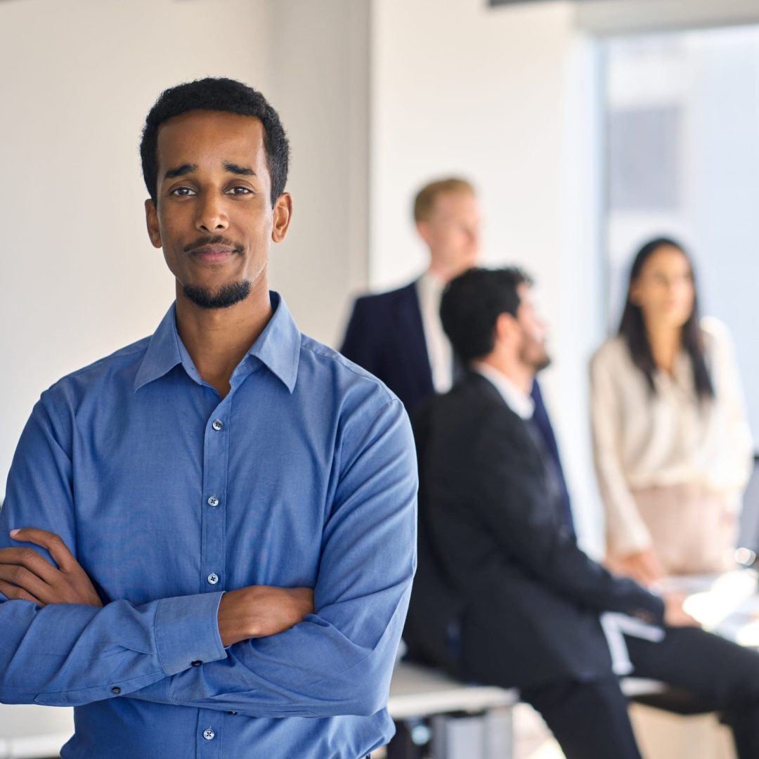 Confident young African American business man worker standing in office looking at camera. Smiling businessman employee corporate leader executive in meeting room with team, portrait.