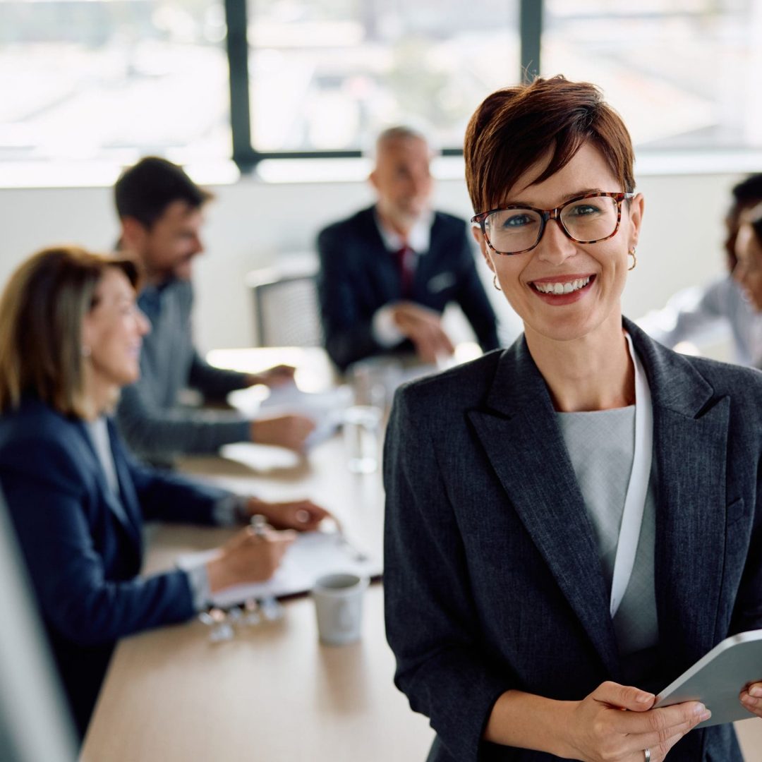 Happy businesswoman with touchpad in meeting room looking at camera. Her colleagues are in the background.