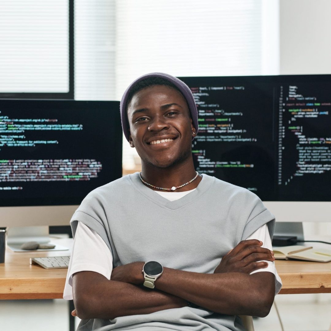 Happy young successful IT engineer in casualwear looking at you while sitting by workplace against computer screens with coded data